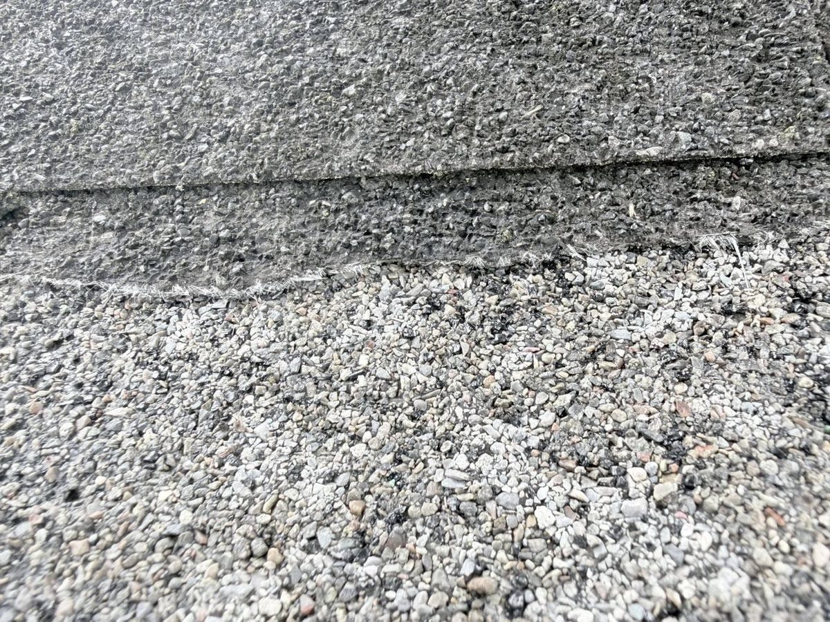 Asphalt shingle worn through to gray asphalt, with fiberglass threads along the edge