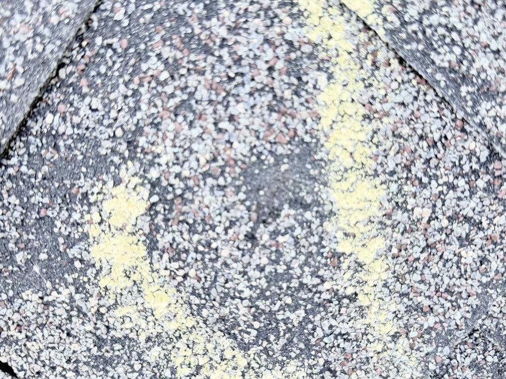 Round dark spot of missing granules on an asphalt shingle, marked with yellow chalk