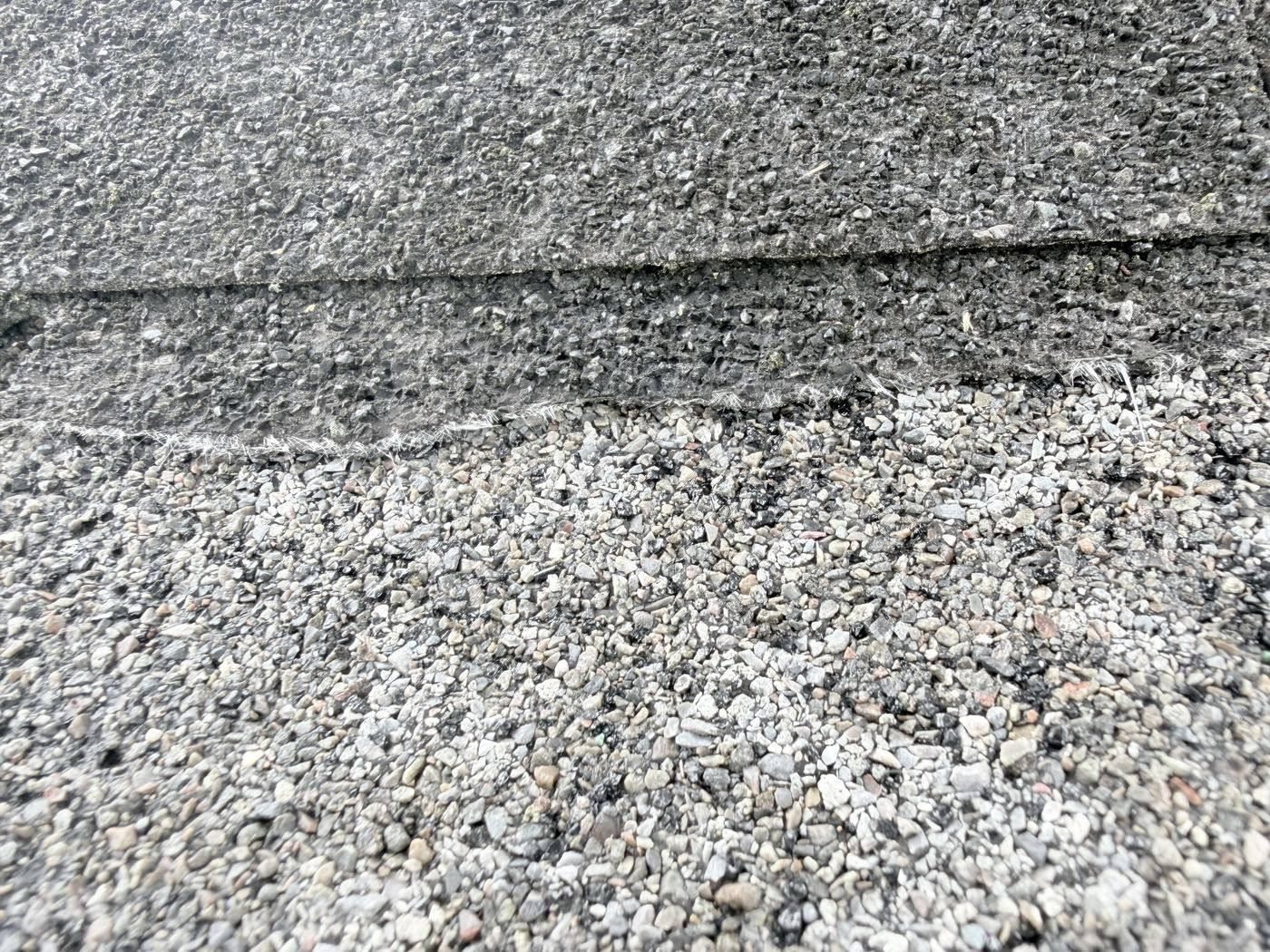 Asphalt shingle worn through to gray asphalt, with fiberglass threads along the edge and loose granules below