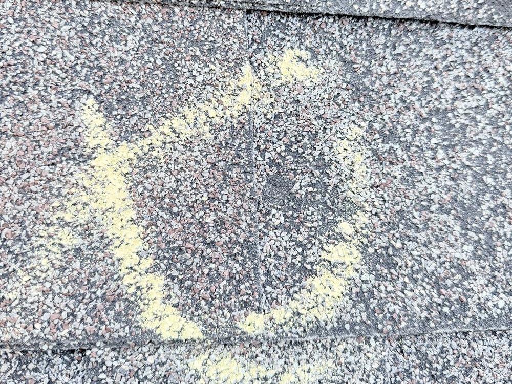 Hail hit on an asphalt shingle circled in yellow chalk, with a dark spot where the granules are missing